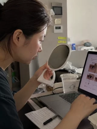 Person comparing different fashion contact lens options on a laptop while checking their eyes in a mirror at home before making a purchase.