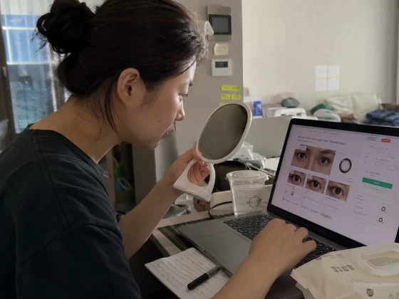 Person comparing different fashion contact lens options on a laptop while checking their eyes in a mirror at home before making a purchase.