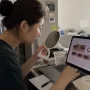 Person comparing different fashion contact lens options on a laptop while checking their eyes in a mirror at home before making a purchase.